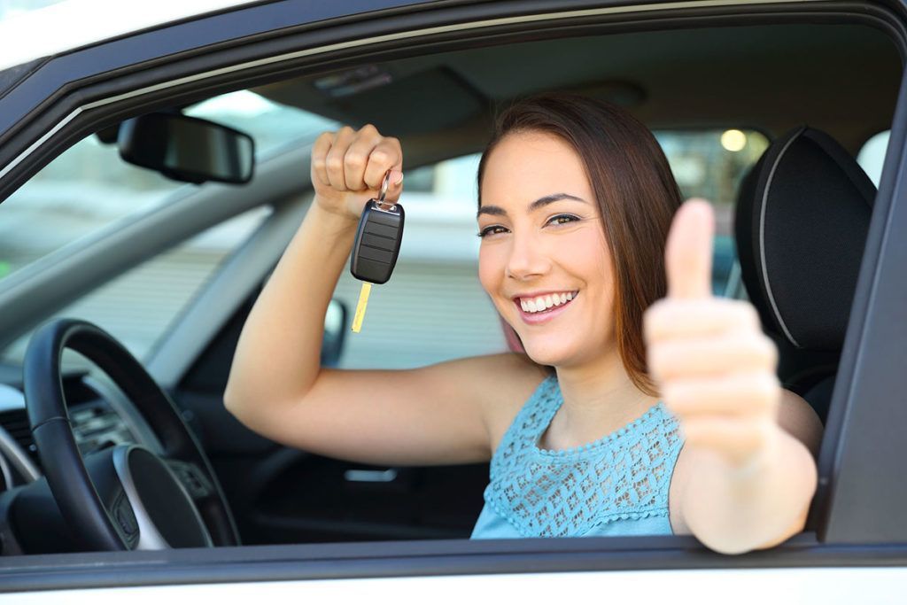 mulher sorrindo dentro do carro olhando para fora da janela segurando a chave do carro com uma mão e com a outra mão apontando o polegar de forma positiva