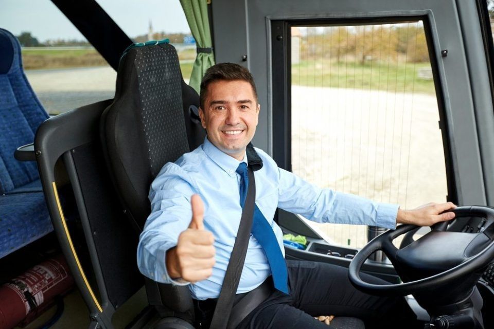 motorista sorrindo sentado em um banco do ônibus com uma mão segurando o volante e a outra apontando para frente com o polegar em movimento de "positivo"
