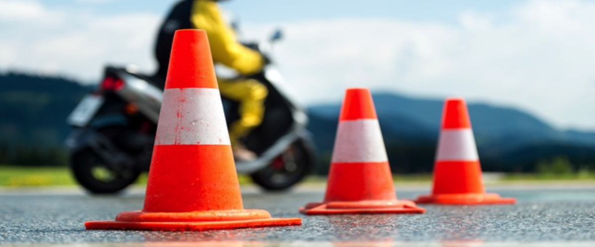 cones de sinalização na cor laranja e branca e no fundo homem andando de moto realizando o teste de baliza em um asfalto